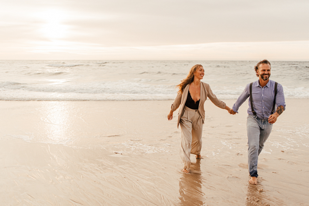 Frau und Mann laufen händchenhalten durch das Wasser am Strand. Beide lachen. Im Hintergrund kommen die Wellen und die Sonne geht unter.