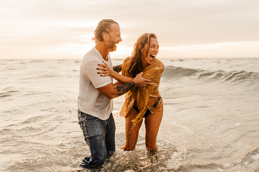 Frau und Mann stehen im Wasser am Strand bei Sonnenuntergang. die Wellen kommen von hinten. Beide lachen und halten sich fest. 