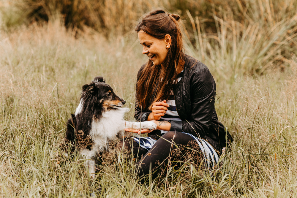 Frau und Hund sitzen im hohen Gras. Hund gibt der Frau die Pfote in die Hand. Frau lacht.