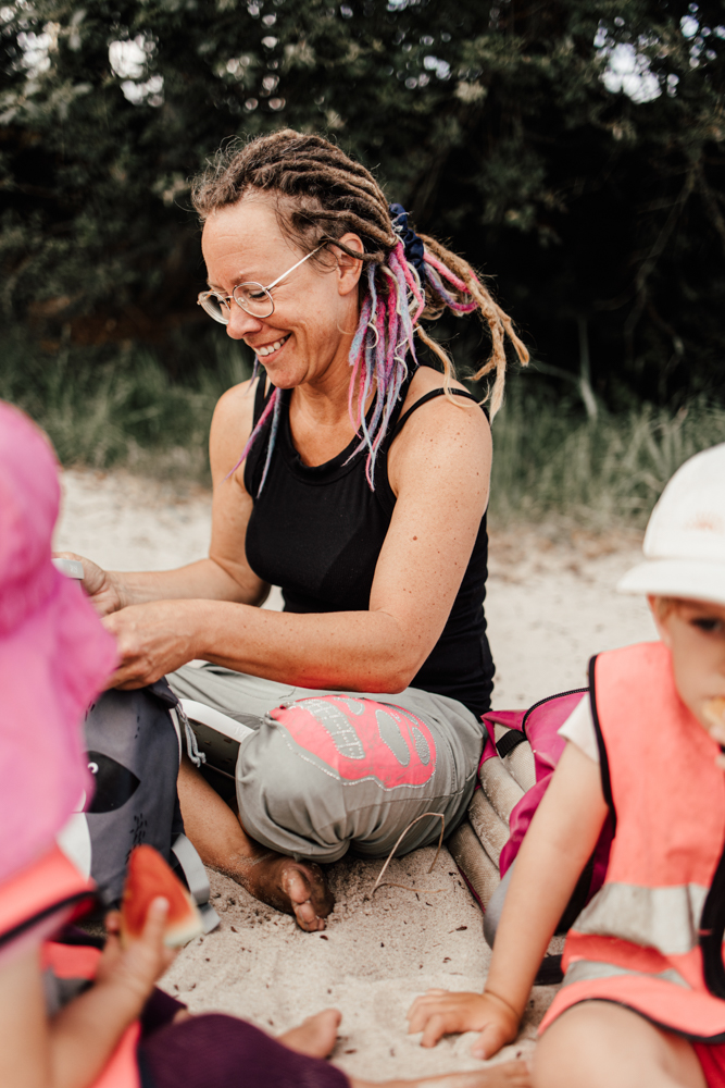 Tagesmutter sitzt im Sand und lächelt. 
Kinder spielen um sie herum