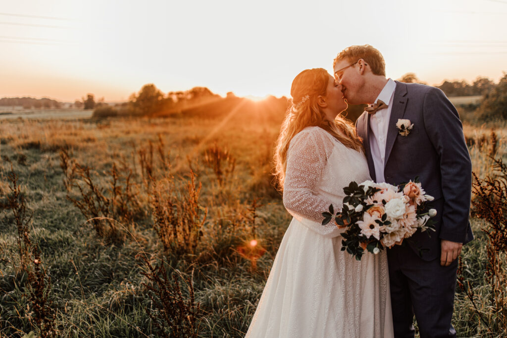 Hochzeitsfotograf und Hochzeitsvideograf in Schleswig-Holstein. Braut und Bräutigam stehen in der freien Natur auf einer Wiese bei Sonnenuntergang.