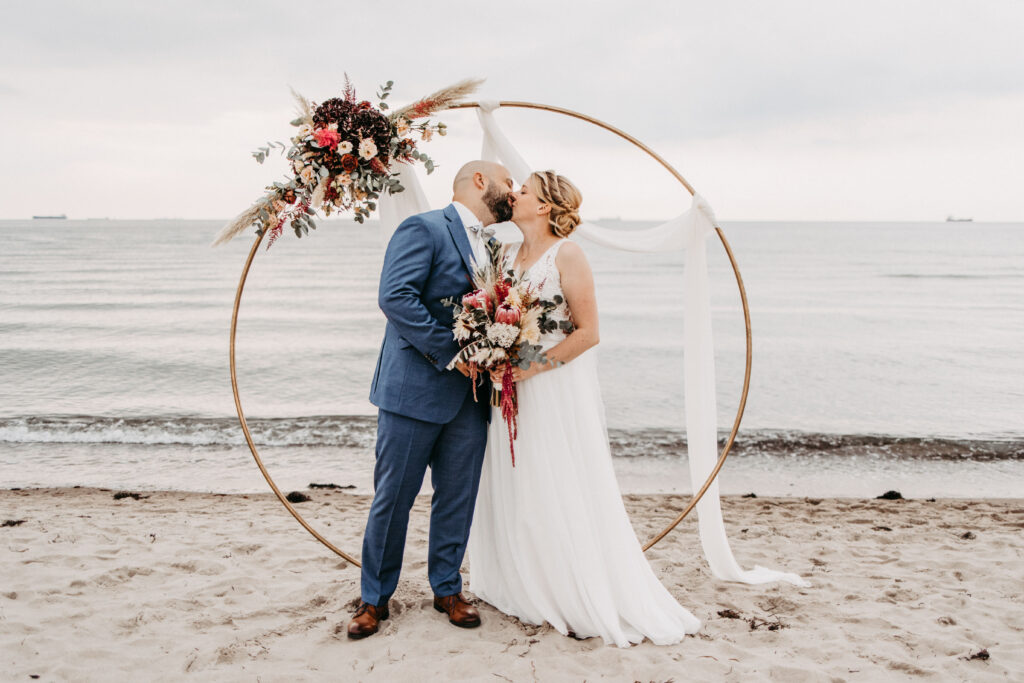 Braut und Bräutigam unter einem dekorativen Ringbogen am Strand von Sylt – einzigartige Hochzeitsfotografie.