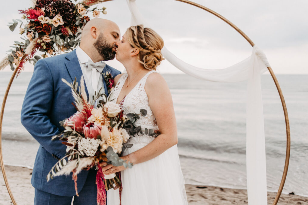 Brautpaar am Strand mit floralem Traubogen – romantischer Moment bei einer Strandhochzeit auf Sylt.