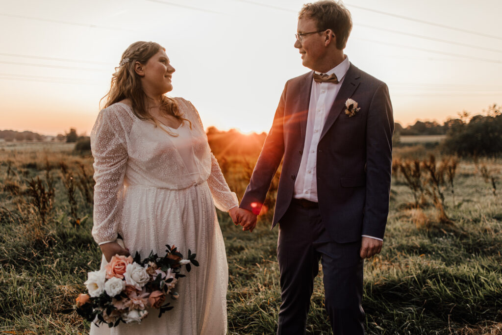 Hochzeitsfotograf und Hochzeitsvideograf in Schleswig-Holstein. Braut und Bräutigam stehen in der freien Natur auf einer Wiese bei Sonnenuntergang.