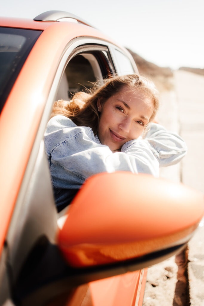 Eine Frau lehnt mit einem strahlenden Lächeln aus dem Fenster eines orangefarbenen Autos. Der Wind spielt mit ihrem Haar, während sie die Weite der Landschaft von Sylt genießt. Ein Moment purer Lebensfreude, perfekt eingefangen für Sylt-Travel-Abenteuer.