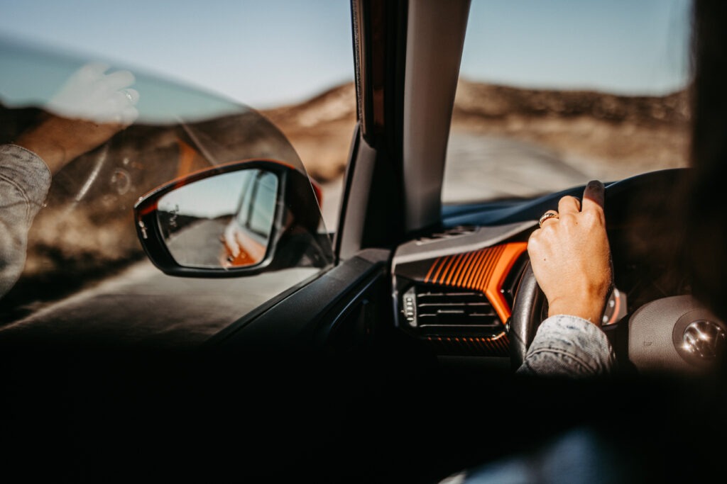 Ein Blick aus dem Inneren eines Autos auf die Straße von Sylt. Die Hand einer Frau greift entspannt das Lenkrad, während der Seitenspiegel die weite Landschaft widerspiegelt. Ein Moment der Freiheit, typisch für das Abenteuer einer Sylt-Travel-Reise.