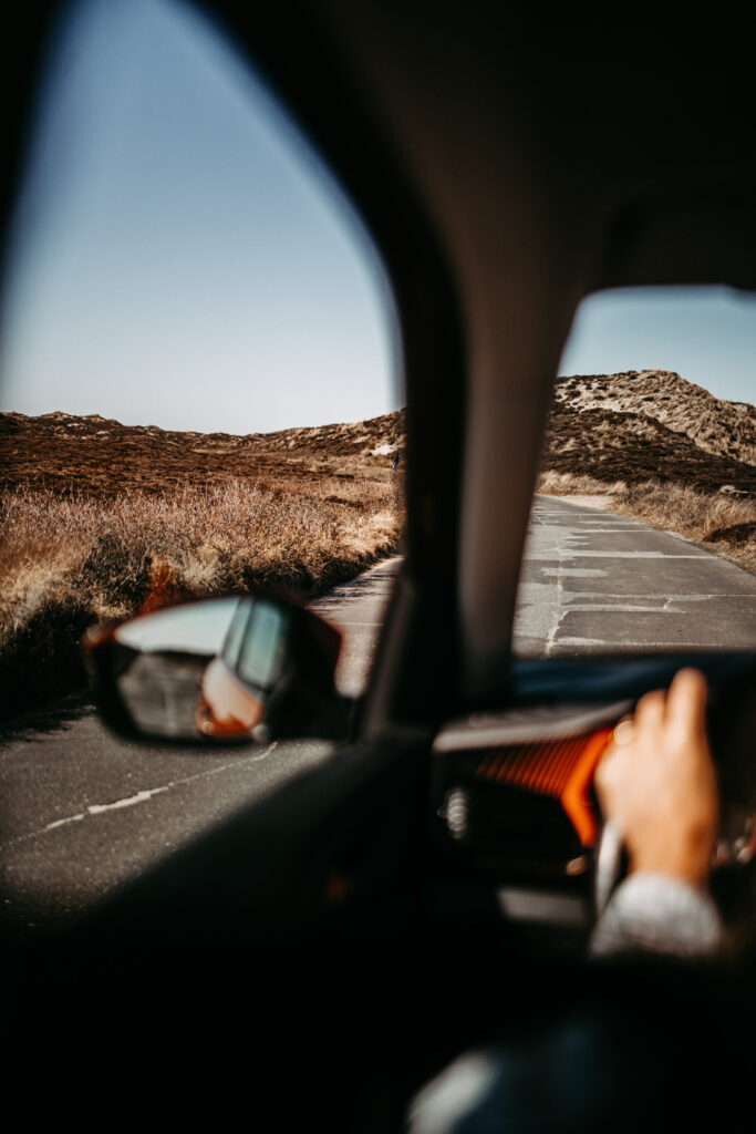 Blick aus dem Innenraum eines Autos, das auf einer weiten, ländlichen Straße auf Sylt fährt. Eine Hand greift das Lenkrad, während der Außenspiegel die offene Landschaft widerspiegelt. Ein Moment der Fahrt, der das Gefühl von Freiheit und Abenteuer auf einem Sylt-Travel-Ausflug vermittelt.