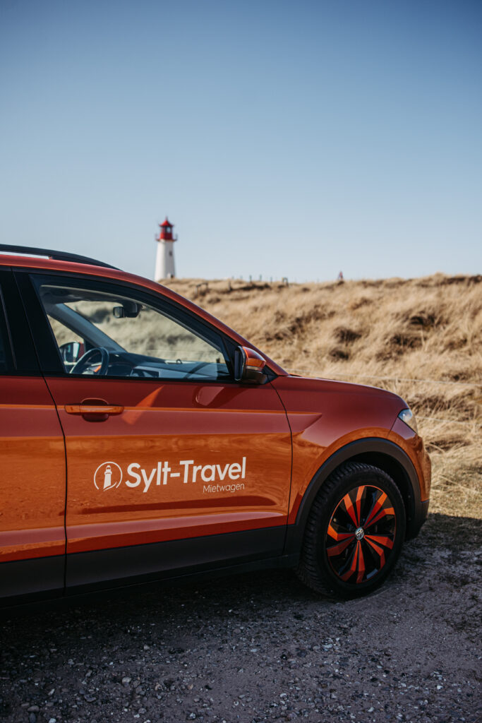 Ein orangefarbenes Auto mit dem Sylt-Travel-Logo fährt auf einer sandigen Strecke, im Hintergrund ist ein Leuchtturm zu sehen. Die weite Landschaft von Sylt und der klare Himmel vermitteln das Gefühl von Freiheit und Abenteuer auf einer Reise.