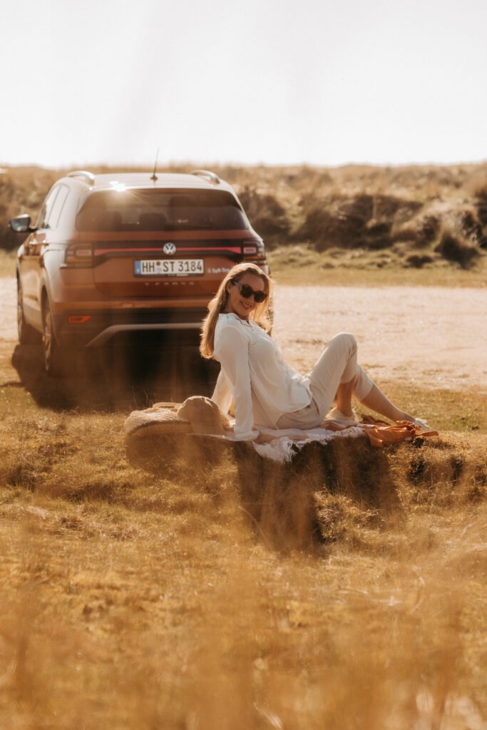 Eine Frau sitzt entspannt im Gras neben einem orangefarbenen Auto, das auf einer weiten Wiese in Sylt geparkt ist. Sie trägt ein weißes Kleid und genießt die Sonne in einer ruhigen Momentaufnahme, die das Gefühl von Freiheit und Entspannung während eines Sylt-Travel-Abenteuers vermittelt.