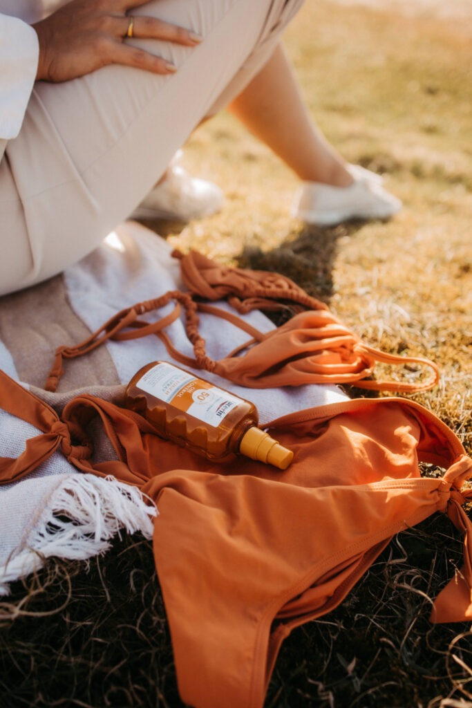 Ein orangefarbenes Strandtuch und eine Sonnencreme-Flasche liegen auf dem Gras, während eine Hand eine entspannte Pose einnimmt. Die warmen Farben und das sommerliche Flair erinnern an unvergessliche Momente bei einem Sylt-Travel-Ausflug.