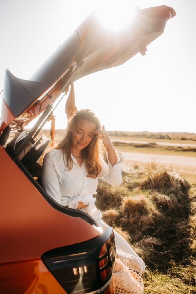 Eine Frau sitzt entspannt im offenen Kofferraum eines Autos auf Sylt. Sie trägt ein weißes Sommerkleid, im Hintergrund ist eine Dünenlandschaft in warmem Sonnenlicht zu sehen. Das Bild strahlt Ruhe und Urlaubsgefühl aus – ein Moment voller Leichtigkeit beim Sylt-Travel.