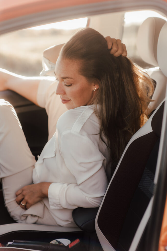Eine Frau sitzt entspannt im Auto, ihre Haare in einer locker-fließenden Bewegung. Sie dreht sich zur Seite, während das warme Licht die Szene erleuchtet. Ein Moment der Ruhe und Vorfreude, der perfekt zu einem Sylt-Travel-Ausflug passt.