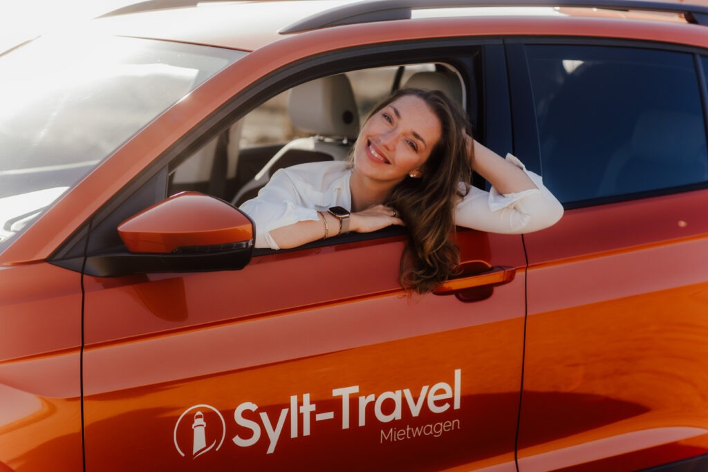 Mit einem Lächeln im Gesicht lehnt sich eine Frau entspannt aus dem Fenster eines leuchtend orangefarbenen Autos, auf dem das Sylt-Travel-Logo zu sehen ist. Die Sonne strahlt in ihr Gesicht – ein perfekter Moment, um die Freiheit und Abenteuerlust auf Sylt zu genießen. Ideal für unvergessliche Sylt-Travel-Momente.
