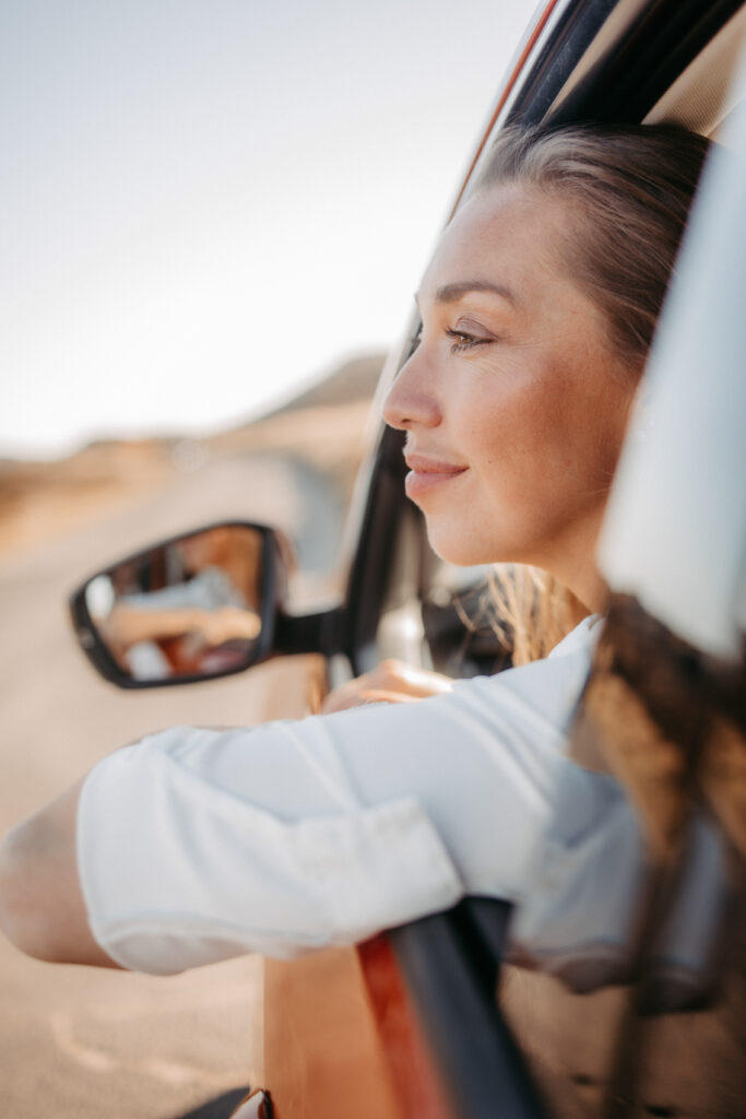 Eine Frau lehnt entspannt aus dem Fenster eines Autos, ihr Gesicht im warmen Sonnenlicht. Der Wind weht sanft durch ihre Haare, während sie mit einem friedlichen Lächeln die Landschaft von Sylt genießt – ein Moment der Ruhe und Freiheit auf einer Sylt-Travel-Reise.