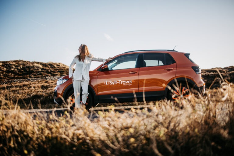 Eine Frau steht neben einem orangefarbenen Auto, das auf einer weiten, offenen Straße auf Sylt parkt. Sie trägt ein weißes Outfit und blickt mit einem Lächeln in die Ferne. Die goldenen Wiesen und der klare Himmel unterstreichen das Gefühl von Freiheit und Abenteuer, typisch für ein Sylt-Travel-Erlebnis.
